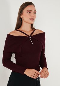 Sieviete, tērpusies bordo ribota adījuma topā ar pleciem atklātu dizainu un sirds formas pogām, kombinēts ar melniem apakšā, pret vienkāršu fona.