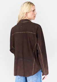 Brun mockaskinnjacka med guldfärgade nitar längs sömmarna, krage och en logotyp på baksidan. Avslappnad passform med sänkta axlar och fickor.