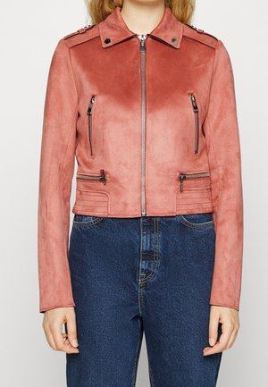 Frau trägt eine kurze pinke Wildlederjacke mit Reißverschluss vorne und vier Reißverschlusstaschen, kombiniert mit hoch taillierten blauen Jeans.