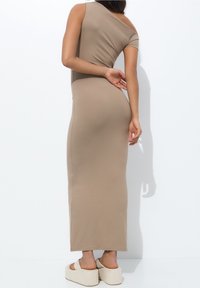Robe maxi moulante marron clair avec un design asymétrique à épaules dénudées. Texture douce avec une silhouette élégante, associée à des sandales à plateforme.