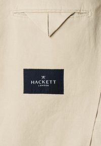 Beiger Stoff mit einer dreieckigen, abgesteppten Klappe über einem schwarzen, rechteckigen Hackett London Stoffetikett.