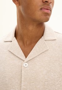 Beige texturerad skjorta i ett mjukt, vävt tyg. Har knäppning fram och en krage med lätt spets. Enkelt mönster utan tryck.