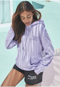 Lila Kapuzenpulli mit Fronttasche und Kordelzügen. Auf der Brust steht: "GENIEßE JEDEN MOMENT DEINES LEBENS." Kombiniert mit schwarzen Shorts, die einen ähnlichen Text haben.