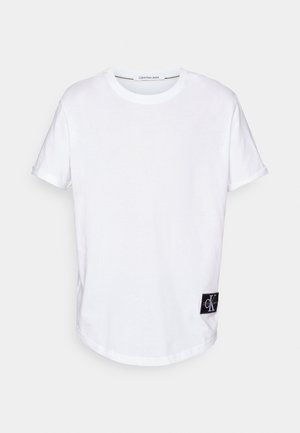 White cotton T-shirt with rolled short sleeves, rounded hem, and a black logo tag featuring "CK" at the bottom left side.