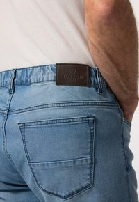 Světle modré džíny mají hladkou strukturu, elastický pas a hnědou koženou nášivku s nápisem "BOSTON PARK" na zadní straně.