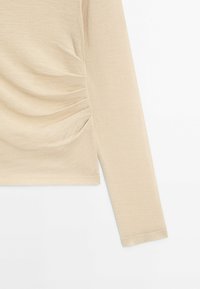 Beige long-sleeve top with soft, smooth texture and gathered side detail. Simple design with a fitted silhouette and regular sleeve length.