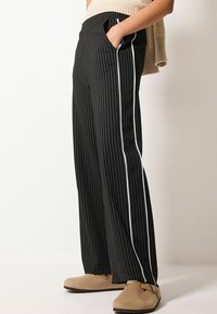 Schwarz gestreifte, weit geschnittene Hose mit weißen Seitenstreifen, gefertigt aus einem glatten Stoff. Verfügt über Seitentaschen und einen lässigen Schnitt.