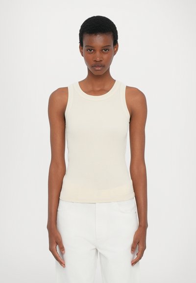 Cream ribbed tank top with a round neckline, fitted shape, and a seamless design, paired with white pants. No notable patterns or accents.