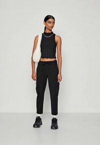 Mlada žena s glatkom repom, nosi bezrukavi crni cropped top, crne cargo hlače, crne tenisice, bijele čarape i bijelu torbu preko ramena.