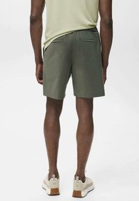 Shorts hommes vert olive avec un tissu lisse, une taille élastique et deux poches arrière. Assortis à des baskets beiges.