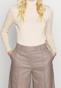 Kvinna iklädd beige polotröja instoppad i hög midja rutig byxor med händerna i fickorna, från midjan till nacken synlig.