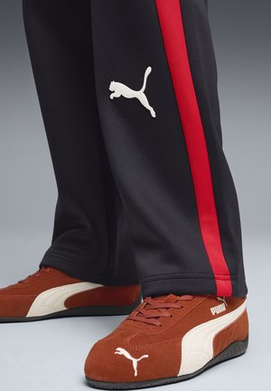 Czarne spodnie dresowe z czerwonymi paskami po bokach i białym logo Puma, zestawione z brązowymi zamszowymi trampkami z białymi akcentami i logo.