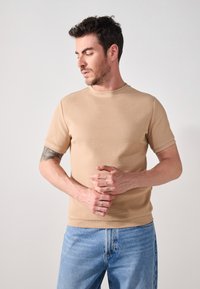 Beige ribbed short-sleeve shirt with a round neck; paired with light blue denim jeans. Notable texture and simple design.