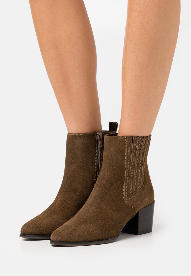 Bottines marron en daim avec talons blocs et fermeture éclair latérale portées sur des jambes nues sur un fond uni.
