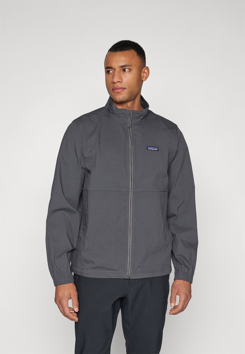 Gray zip-up jacket with a high collar, features elastic cuffs and a simple design. Logo patch displayed on the left chest area.