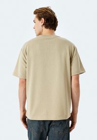 T-shirt di cotone beige con maniche corte, vestibilità rilassata e una texture sottile, indossato con jeans in denim blu, visto da dietro.