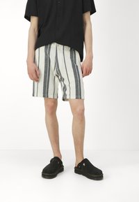 Man i svart kortärmad skjorta med knappar, vita shorts med svarta vertikala ränder och svarta slip-on skor som står på vit golv.