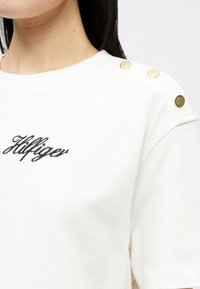 Κοντινό πλάνο ενός λευκού πουκαμίσου με μαύρη κεντημένη επιγραφή "Hilfiger" και τρία χρυσά κουμπιά στον ώμο, φορεμένο από ένα άτομο με σκούρα μαλλιά.