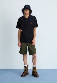 Polo noir avec un logo rouge, short vert olive et bottes à lacets marron avec des accents jaunes. Des chaussettes blanches montantes complètent la tenue.