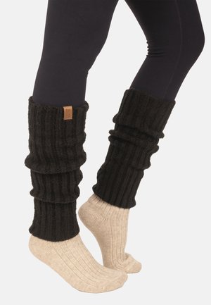 Beine in schwarzen Leggings, beige gerippte Socken und lockere, schwarze gerippte Strick-Beinwärmer, einer mit einem kleinen braunen Etikett.