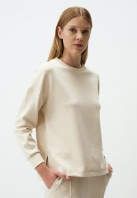 Felpa beige realizzata in materiale morbido, con scollatura a girocollo, maniche lunghe e spacchi laterali per facilitare i movimenti.