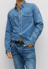 Camisa de denim azul claro con botones al frente, dos bolsillos en el pecho y un cuello clásico. Usada con jeans azules a juego y un cinturón negro.