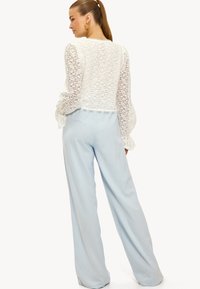 Top en dentelle blanche à manches longues, bords festonnés, associé à un pantalon large bleu clair, présentant une texture lisse et une coupe décontractée.