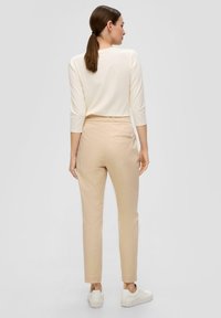 Pantalon beige fuselé avec une texture lisse, présentant une poche arrière, associé à un haut à manches longues crème et des baskets blanches.