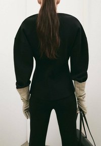 Čierny štruktúrovaný top so širokými rukávmi v kombinácii s dlhými krémovými rukavicami. Oblečenie má priliehavú siluetu na svetlom pozadí.