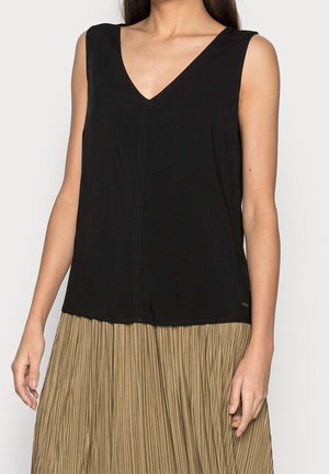 Kvinde iført sort ærmeløs V-hals top kombineret med en højtaljet beige plisseret nederdel, stående foran en ensfarvet baggrund.