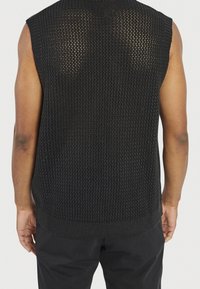 Chaleco de punto sin mangas negro con un patrón de malla texturizada, dobladillo acanalado y amplios brazos, que muestra un diseño casual sin cuello.