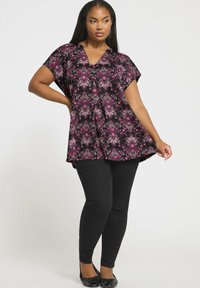 Kobieta nosząca czarne legginsy i czarną tunikę z dekoltem w serek w różowe kwiatowe wzory, pozująca z jedną ręką na biodrze, a drugą trzymająca dół tuniki.