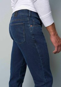 Jean en denim bleu à coupe slim, avec des poches arrière, des coutures discrètes et une étiquette de taille noire. Texture lisse et design classique.