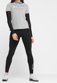 Camiseta de algodón gris con el logo "Champion" en blanco, combinada con leggings negros que tienen un logo blanco en el lateral. Modelo lleva zapatillas deportivas grises.