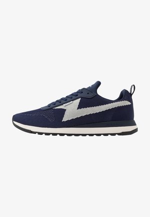 Chaussure de sport bleu marine avec bande blanche en zigzag sur le côté, tige tricotée, fermeture à lacets, semelle amortie et languette à l'arrière.
