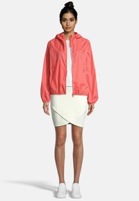 Giacca leggera corallo con cappuccio e polsini elastici, abbinata a una camicia bianca aderente e a una gonna a portafoglio color crema. Completata con sneaker bianche.