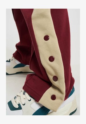 Joggers color burdeos con un panel lateral beige, que cuentan con tres botones a presión en color burgundy. Combinados con zapatillas blancas y verde azulado. Textura de tela suave.