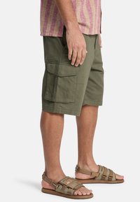 Shorts cargo vert olive avec plusieurs poches, coupe droite et en coton. Associés à des sandales marron à lanières.