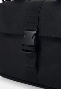 Svart vikbar messengerbag med yta av texturerat tyg, utrustad med en plastspänne och justerbar nylonrem för säker stängning.