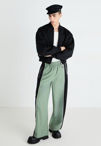 Veste bomber noire, t-shirt blanc, pantalon de sport vert avec panneaux latéraux noirs et rayures blanches, casquette noire, chaussures noires épaisses.