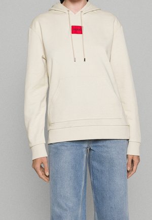 Beige hoodie med en framficka, med en röd rektangulär patch med "HUGO" text. Den har en dragsko på huvan och en avslappnad passform.