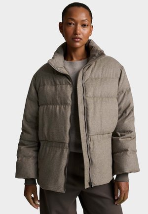 Donna con una giacca imbottita beige con collo alto, in piedi davanti a uno sfondo chiaro e uniforme, rivolta in avanti.