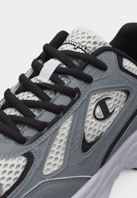 Κοντινή λήψη ενός γκρι και λευκού αθλητικού παπουτσιού με δίχτυ, με μαύρα κορδόνια και το λογότυπο Champion στο πλάι και στη γλώσσα του παπουτσιού.