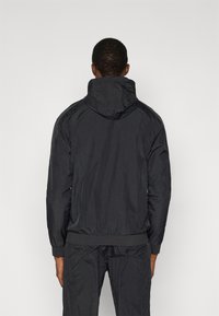 Μαύρο ελαφρύ μπουφάν με κουκούλα, με ελαστικές μανσέτες και κάτω μέρος, κατασκευασμένο από ζαρωμένο ύφασμα, εμφανιζόμενο από πίσω.