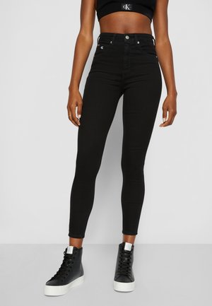 Femme portant un jean skinny taille haute noir, un top court noir et des baskets montantes noires avec semelles blanches, debout devant un fond gris.