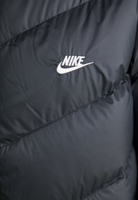 Sort puffer-jakke med et diagonalt quiltet mønster og et hvidt Nike-logo, der er tydeligt placeret på brystområdet. Glat, blankt stof.