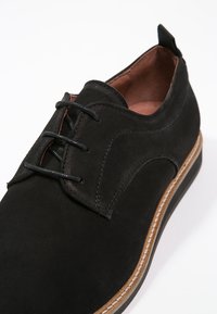 Scarpe stringate in suede nero con suola cucita marrone e linguetta sul tallone, presentate su uno sfondo bianco.