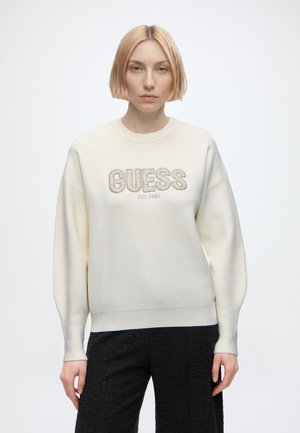 Jeune femme aux cheveux blonds courts portant un sweat-shirt crème GUESS et un pantalon noir, debout devant un fond blanc uni.