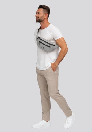 Man in wit T-shirt, beige broek en witte sneakers staat met een hand in de zak, met een grijze schoudertas op de borst.