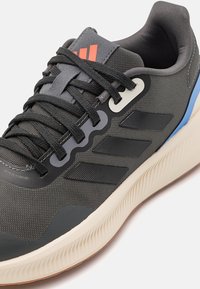 Zapatilla deportiva gris con cordones negros, parte superior de malla texturizada, tres rayas negras, suela blanca y detalles rojos en la lengüeta.